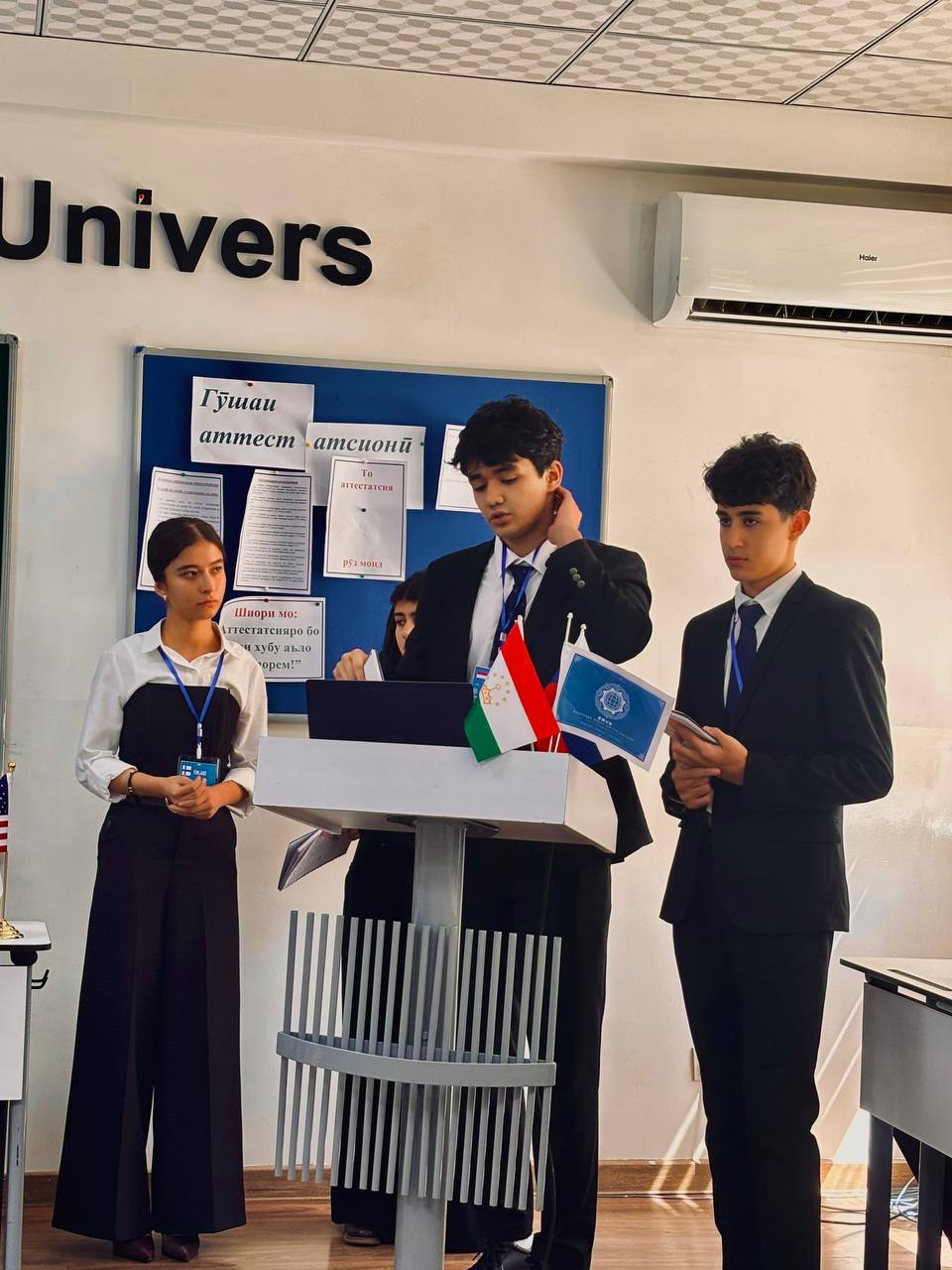 Delegates in formal dress standing at a podium with Tajik and DMUN table flags during committee.
